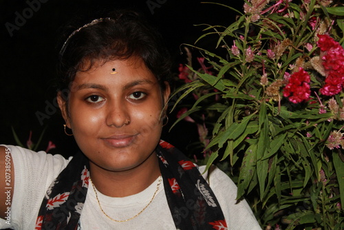 Portrait of a young Indian woman posing beside flowering plants in an outdoor garden at night. The image captures natural beauty, confidence, and contemporary Indian lifestyle, with vibrant flowers