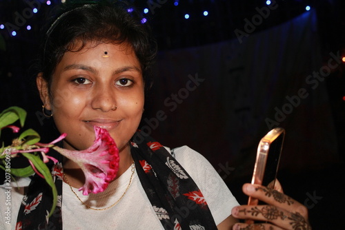 Authentic portrait of a young Indian woman taking a selfie with a smartphone while holding a pink flower under decorative lights at night. The image captures a cheerful expression, natural beauty