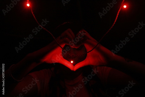 Creative low-light image of hands forming a heart shape around a glowing red LED light against a black background. The warm red illumination creates a dramatic and romantic atmosphere