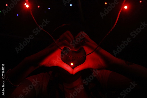 Creative low-light image of hands forming a heart shape around a glowing red LED light against a black background. The warm red illumination creates a dramatic and romantic atmosphere