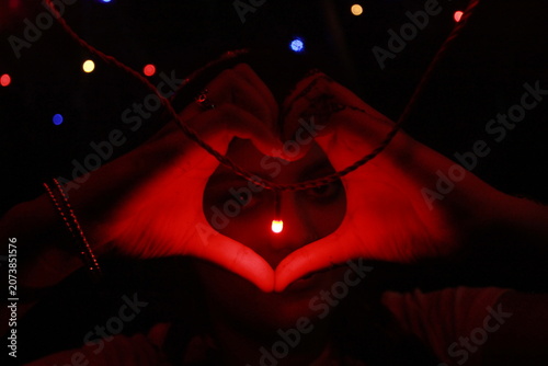 Creative low-light image of hands forming a heart shape around a glowing red LED light against a black background. The warm red illumination creates a dramatic and romantic atmosphere