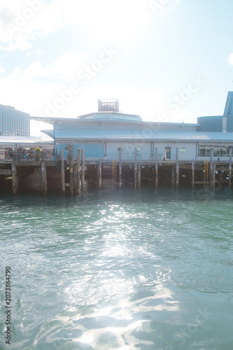 Auckland, New Zealand: vertical bright sunburst lens flare over emerald ocean water next to historic wooden pier piles at downtown ferry terminal.