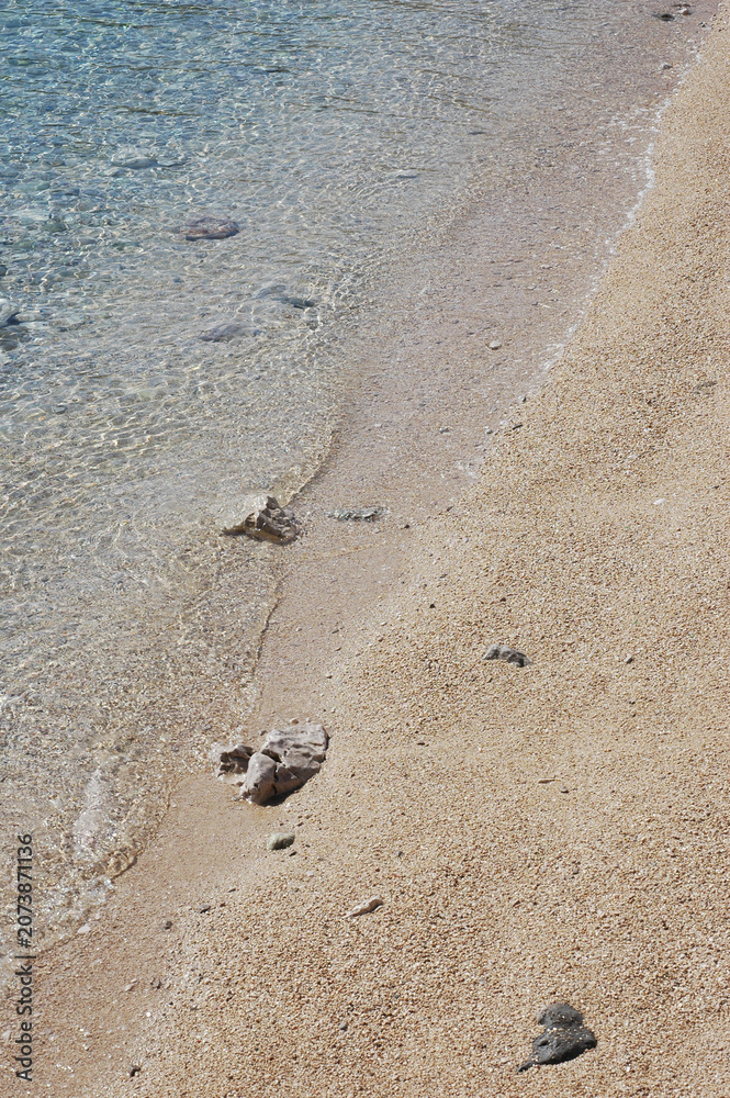 Obraz premium A high angle view of clear turquoise sea water gently washing onto a clean golden sand and pebble beach during a bright sunny day.