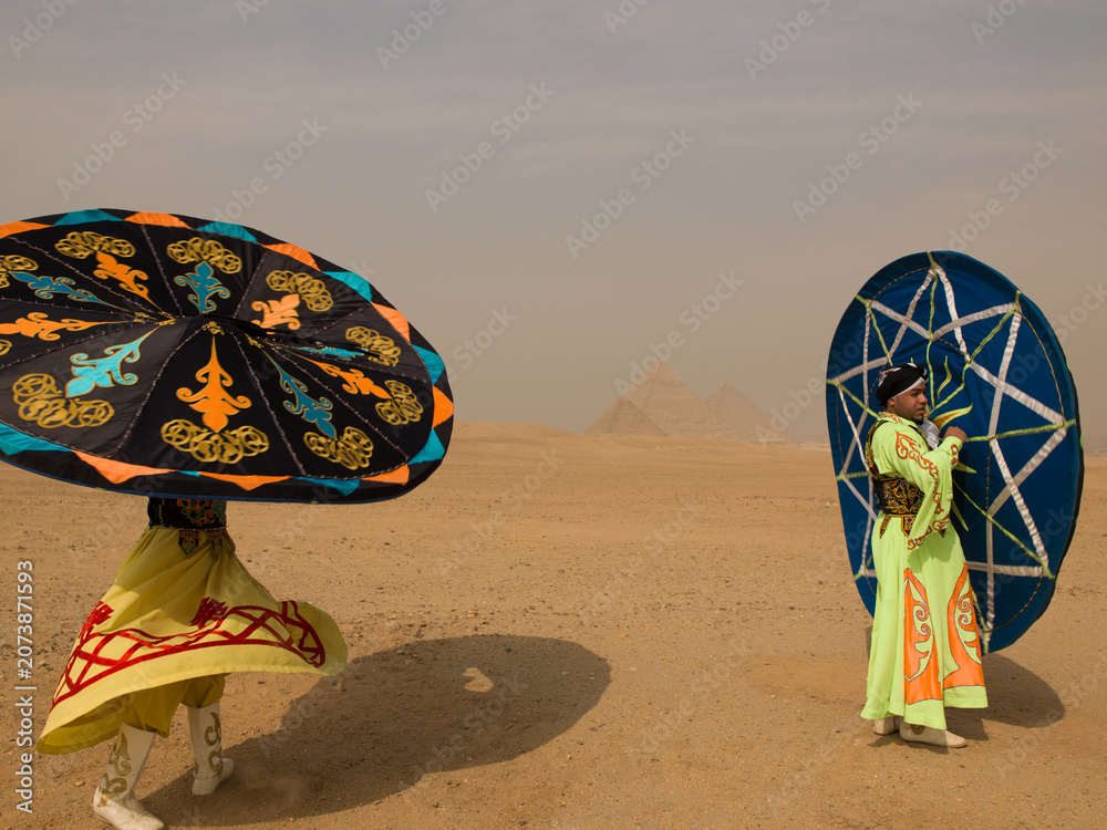 Fototapeta premium Two Egyptian tanoura dancers joyfully spinning vivid folkloric skirts overhead in the desert before the Giza pyramids ideal for cultural tourism and