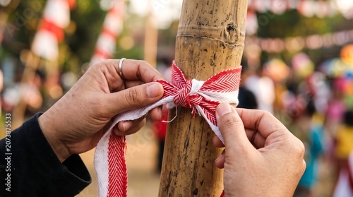 Person tying red and white decorative ribbon onto bamboo structure outdoors.