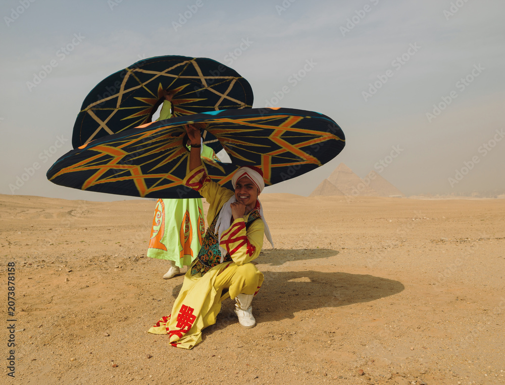 Fototapeta premium Festive Egyptian tanoura dancer spins a colorful patterned skirt overhead and smiles while kneeling in the sandy desert before the ancient Giza