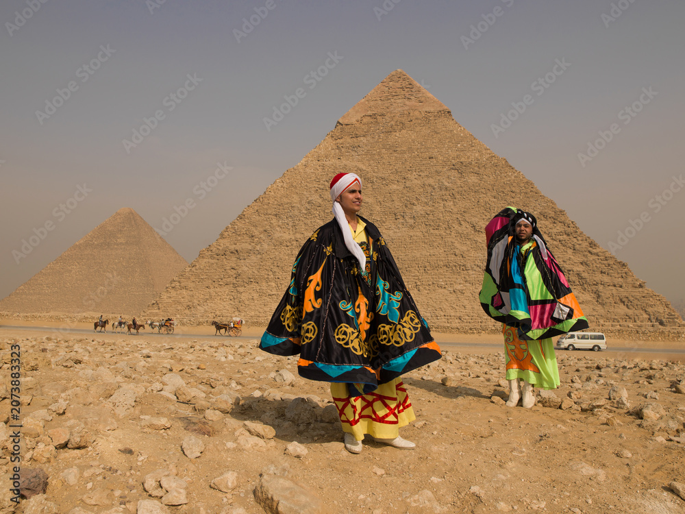 Fototapeta premium Two Egyptian tanoura folk performers in vibrant colorful spinning dance robes standing proudly before the Giza pyramids under bright sun ideal for