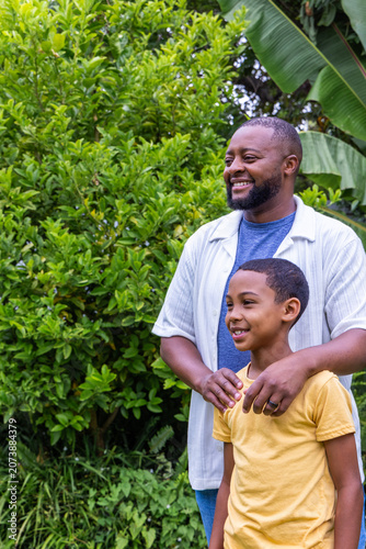 African American father and son standing in backyard garden among banana leaves, hands on shoulders