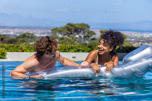 Diverse couple playing on silver inflatable lounger at pool, wearing trunks, bikini and sunglasses