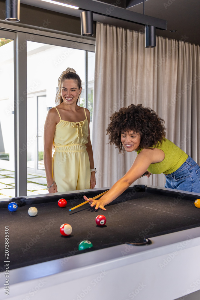 Naklejka premium Diverse female friends leaning and shooting cue at black-felt pool table, wearing colorful clothes