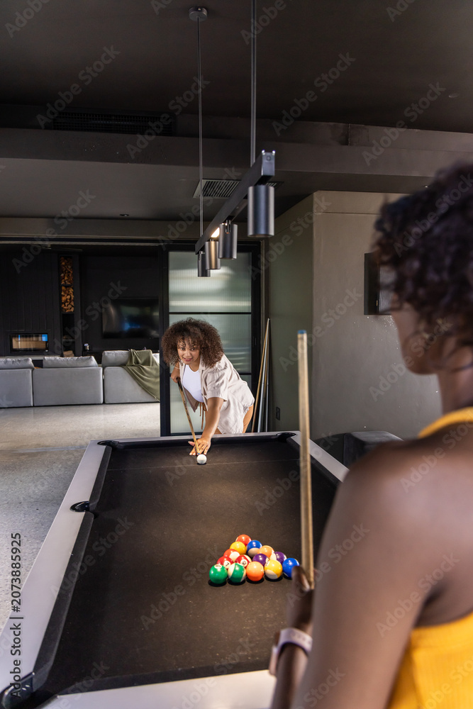 Naklejka premium Diverse female friends playing pool in modern game room with black felt table, racked balls, cues