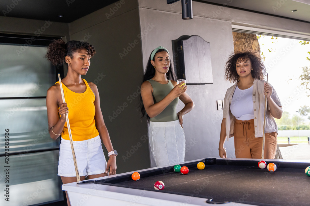Naklejka premium Diverse female friends playing pool in covered game room at black-felt table with cues, balls