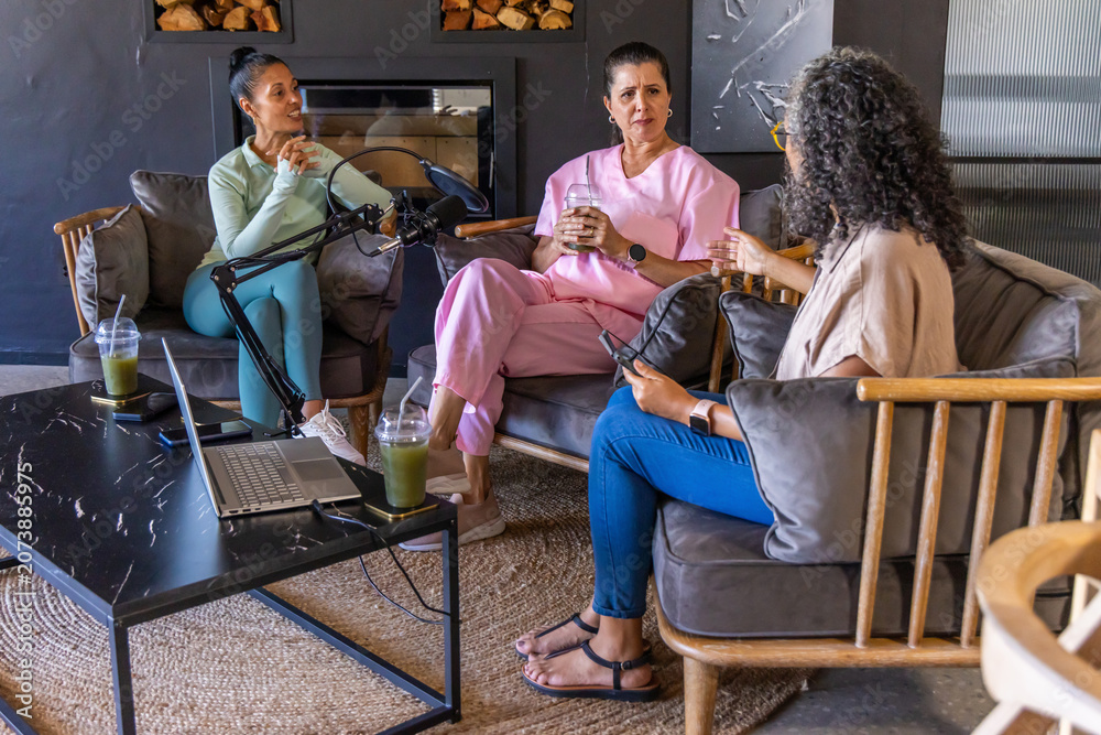 Obraz premium Diverse female friends recording on boom mics in living room, sipping green drinks in lounge wear