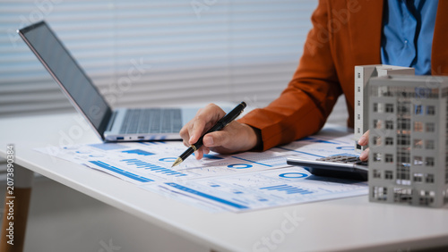 Business professional reviewing financial charts at a desk while holding a small building model, suggesting real estate investment, planning, and market analysis.