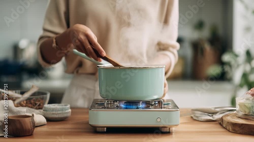 Woman cooking a healthy meal in a modern kitchen, stirring a pot on a stylish gas stove with fresh ingredients nearby. Home cooking and culinary lifestyle concept