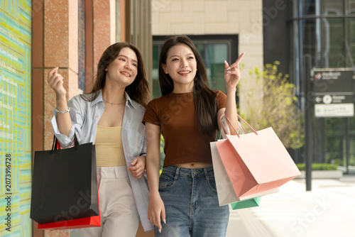 Young two girls enjoys shopping in city. Beautiful Asian women with colorful shopping bags walking in downtown, pointing something. Happy close friends walk through the mall together, shop in stores.