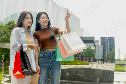Young two girls enjoys shopping in city. Beautiful Asian women with colorful shopping bags walking in downtown, pointing something. Happy close friends walk through the mall together, shop in stores.