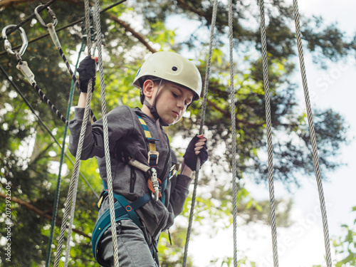 A horizontal medium shot of a focused young boy wearing a safety helmet and harness holding onto vertical cables as he navigates a high ropes challenge course in an outdoor adventure park