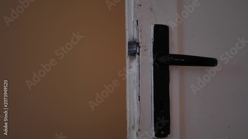 Close-up video of a vintage black door handle on an old white wooden door gently opening to a dimly lit room.