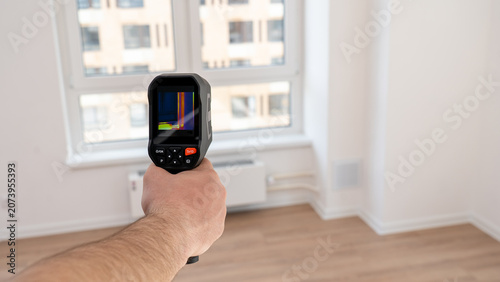 Hand holds thermal imaging device. Screen shows heat patterns in color. Room has light wood floors and white walls. Window reveals city buildings outside. Check for energy leaks or insulation problems