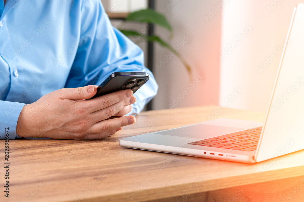 Fototapeta premium Person in light blue shirt holds smartphone. Open laptop rests on wooden table. Soft light creates a calm workspace. This scene suggests focused work. Perfect for business or digital concepts
