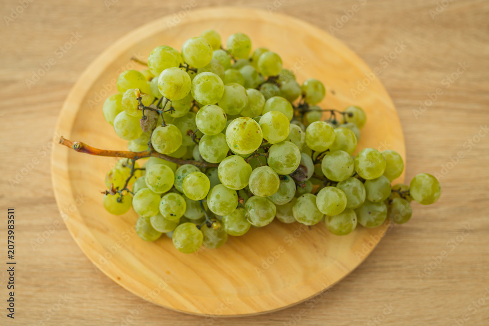 Fototapeta premium Fresh green grapes on a round wooden plate, shot from above on a light wood surface. Perfect for healthy eating and organic food concepts