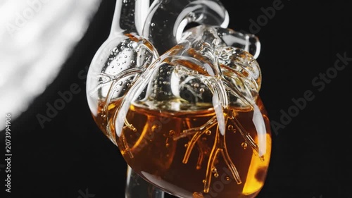 Heart model constructed from translucent glass glows with golden liquid inside a dark laboratory environment.