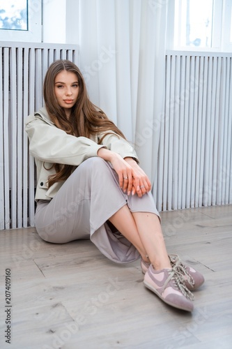 Young woman with long hair seated on floor beside radiator in bright modern interior, wearing neutral outfit and sneakers, relaxed pose with hands clasped and soft natural light