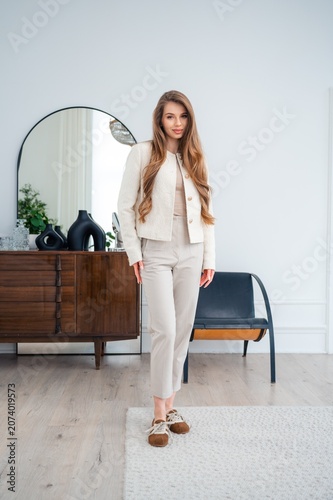 Young woman in neutral outfit standing inside bright modern living room beside wooden sideboard, round mirror, soft natural light, minimalist home decor, calm elegant lifestyle scene