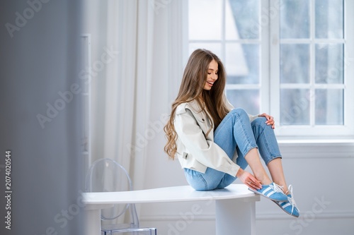 Smiling young woman seated on white table beside bright window, lifting knee to fasten blue sneaker while wearing light jacket and jeans in modern home interior