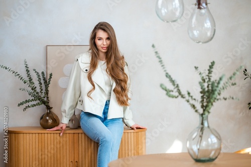Young woman with long wavy hair seated on rounded wooden cabinet in bright minimalist interior, wearing light cream jacket and blue jeans, calm lifestyle portrait
