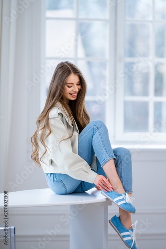Smiling young woman indoors adjusting blue sneaker while seated on raised table beside bright window, casual denim outfit, soft natural light, modern lifestyle portrait