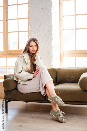 Young woman seated on olive sofa beside bright wooden window, wearing light trench coat, neutral skirt, and green lace-up sneakers, posing with relaxed hand-touch cheek expression