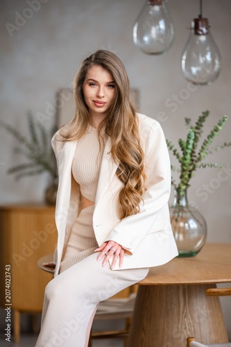 Confident portrait of young woman with long wavy light brown hair wearing cream dress and matching blazer, seated indoors on wooden table edge in softly lit modern interior