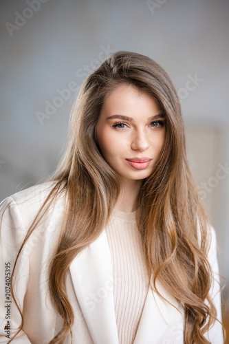 Confident young woman portrait with long brown hair, soft makeup, light blazer, ribbed sweater and direct gaze in softly lit indoor setting, conveying calm modern professionalism