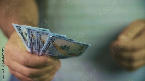 A close-up of a man's hands is visible, holding several hundred-dollar bills with a fan.