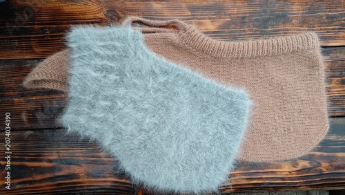 Horizontal flat lay of a fluffy light grey angora knitted hood over a beige wool hood. Handmade winter balaclavas on a dark rustic wooden surface. Cozy accessories
