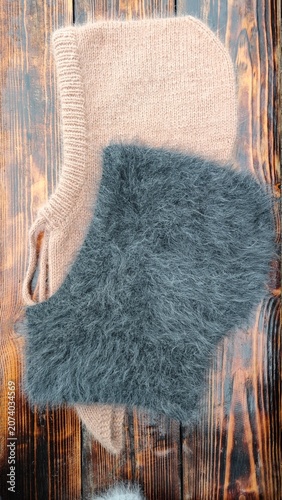Vertical flat lay of two handmade knitted winter hoods on a dark rustic wooden bench. Fluffy charcoal grey mohair hood on top of a beige wool balaclava. Cozy accessories