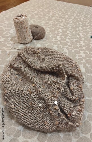 Beige knitted scarf with gold sequins on patterned fabric. Handmade winter accessory with yarn ball and thread spool in background