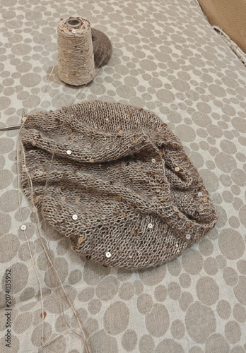 Partially knitted beige fabric with gold sequins on a patterned surface. Handcraft hobby project with yarn spool and knitting needles