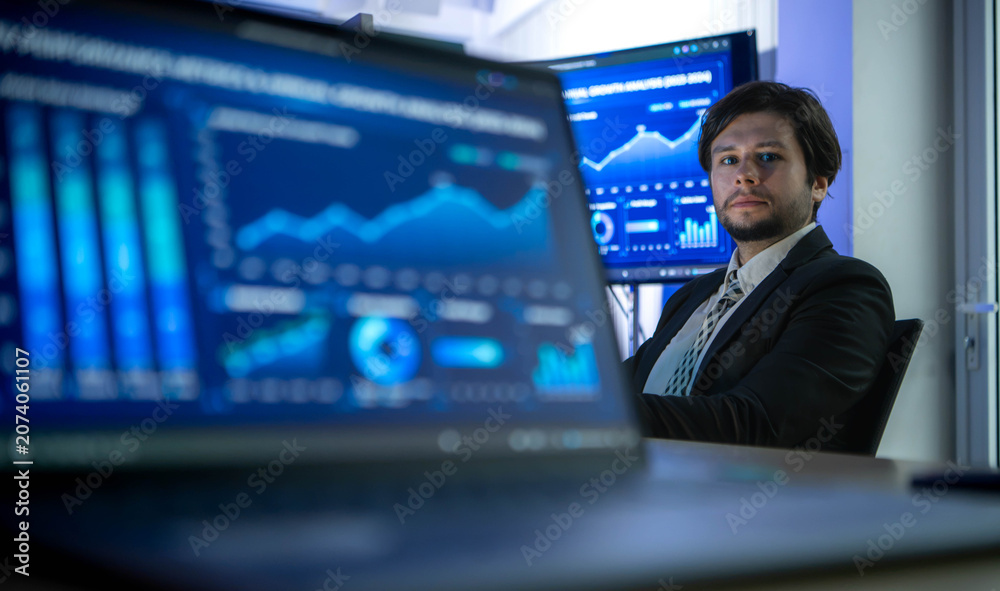 Fototapeta premium Software developer working at a computer while colleagues present business analytics in the background