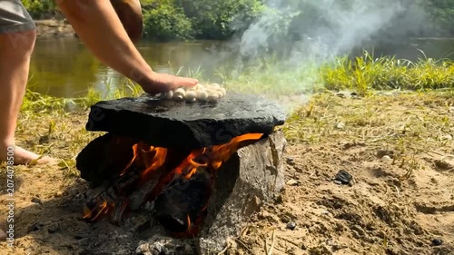 An outdoor scene shows someone placing mushrooms on a hot stone above flames and cutting an eggplant into slices to cook in a natural setting by the river bank in the warm summer daylight hours
