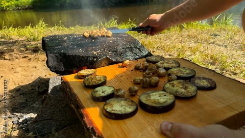 A person transfers grilled mushrooms, eggplant slices, and a large tomahawk steak from a hot stone onto a rustic wooden serving board in a natural riverside setting during the bright daylight hours
