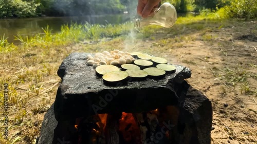 A person seasons mushrooms and eggplant slices with oil and spices while grilling them on a large flat stone over a campfire in a serene riverside environment during the bright daylight hours

