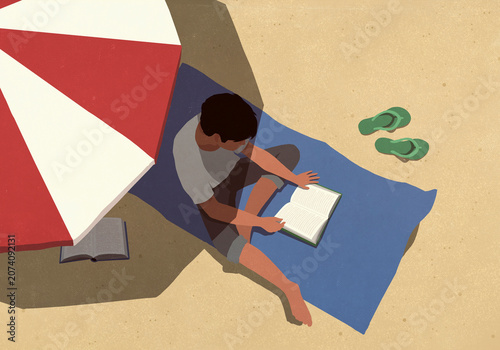 High angle view young man reading book in shade under striped umbrella on sunny summer beach