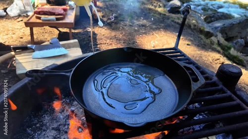 Two seasoned beef steaks are placed into a hot cast iron skillet containing oil and seared over open flames at a campsite to prepare a gourmet meal in the wilderness during an outdoor trip