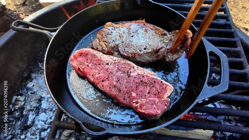 Two seasoned beef steaks are placed into a hot cast iron skillet containing oil and seared over open flames at a campsite to prepare a gourmet meal in the wilderness during an outdoor trip