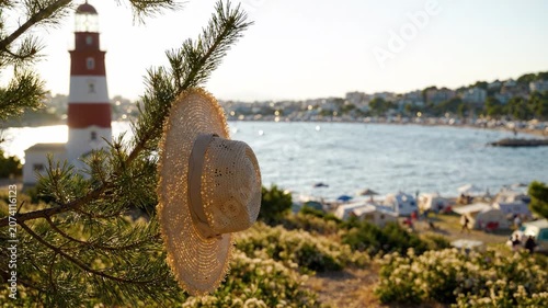 A Scenic Seaside View Featuring a Straw Hat Hanging from a Pine Tree with a Beautiful Lighthouse and Beach in the Background During a Sunny Day