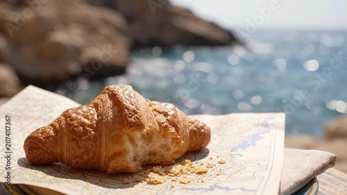 A delicate croissant sits invitingly on a map against a beautiful seaside backdrop, capturing the blend of fine pastry artistry and nature's serene beauty