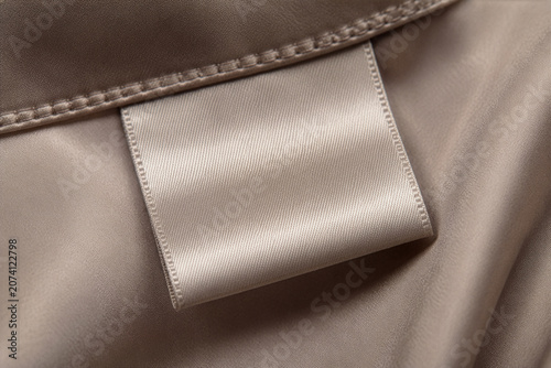 Blank beige satin clothing label mockup sewn into luxury fabric. Premium garment tag close up for fashion brand logo presentation, apparel design and textile branding.